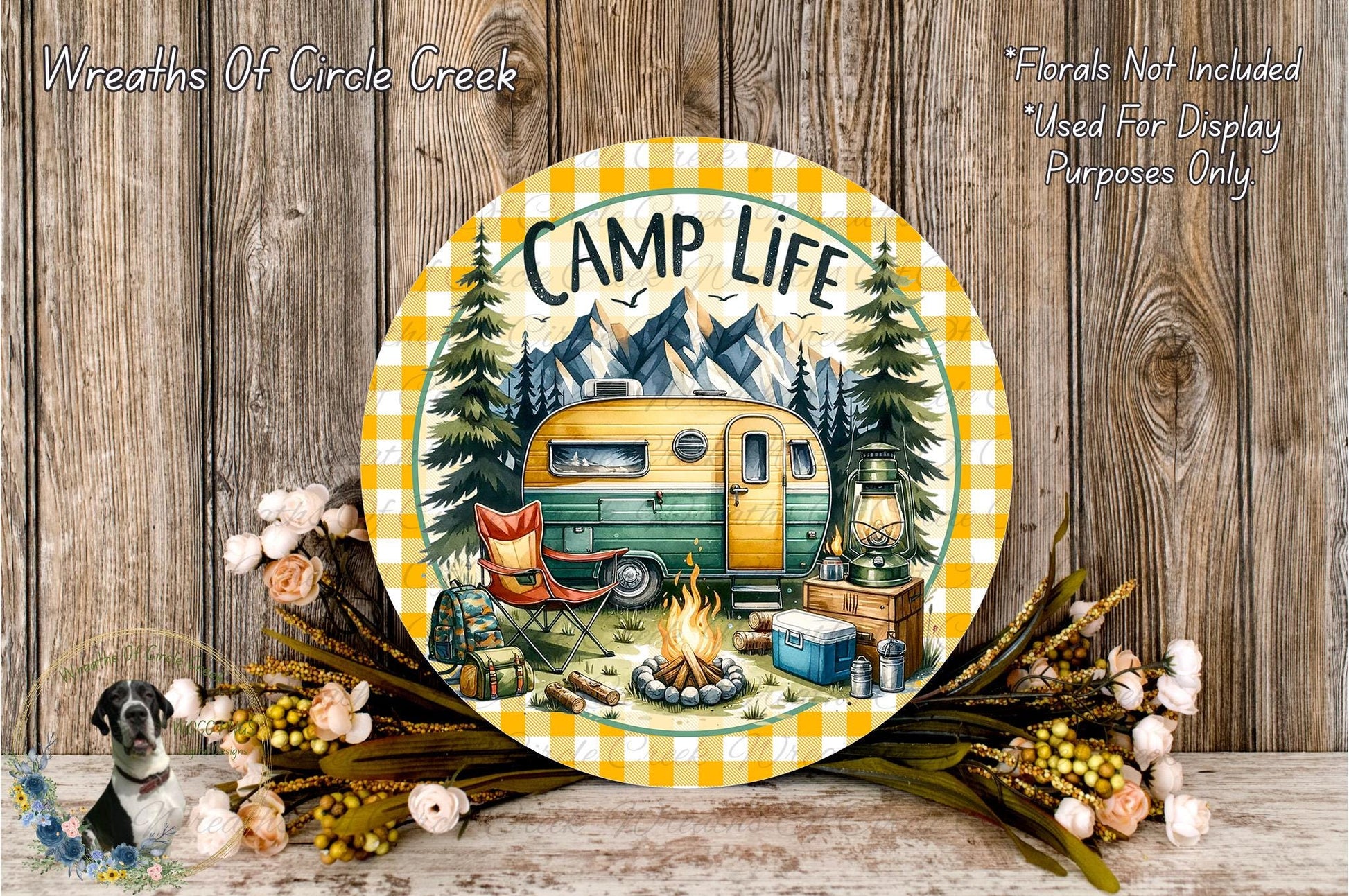 A round wooden plaque with a camping scene, including a camper van, campfire, and camping gear, surrounded by flowers and greenery.
