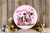 A round, decorative plate featuring a cow wearing a pink hat and boots, surrounded by pink flowers and greenery, with the word "Howdy" written on it.