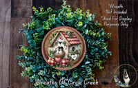 A wreath with a house and birds on it, surrounded by greenery, is displayed on a wooden surface.