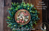 A wreath with a house and birds on it, surrounded by greenery, is displayed on a wooden surface.
