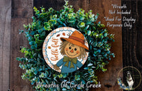 a wreath with a scarecrow design, surrounded by greenery. The wreath is placed on a wooden surface, and there is a small dog in the bottom right corner of the image.