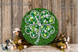 A large, ornate, and colorful four-leaf clover-shaped wreath made of green glitter and rhinestones, placed on a wooden surface with some flowers and greenery.