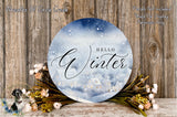 a round sign with the word "Hello Winter" written on it, surrounded by a wreath of flowers and a snowy landscape. The sign is placed on a wooden surface, and there is a small dog in the background.