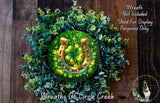 A wreath made of green leaves and a horseshoe with a four-leaf clover on it, placed on a wooden surface.