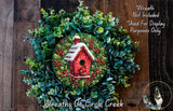A wreath made of green leaves and red berries, with a small red birdhouse in the center, is displayed on a wooden surface.