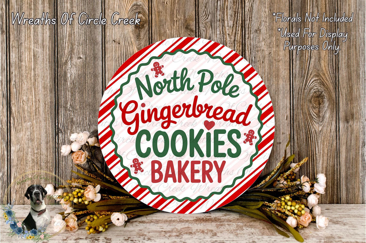 A round sign with the text "North Pole Gingerbread Cookies Bakery" displayed prominently, surrounded by a wreath of flowers and a wooden background.