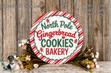 A round sign with the text "North Pole Gingerbread Cookies Bakery" displayed prominently, surrounded by a wreath of flowers and a wooden background.