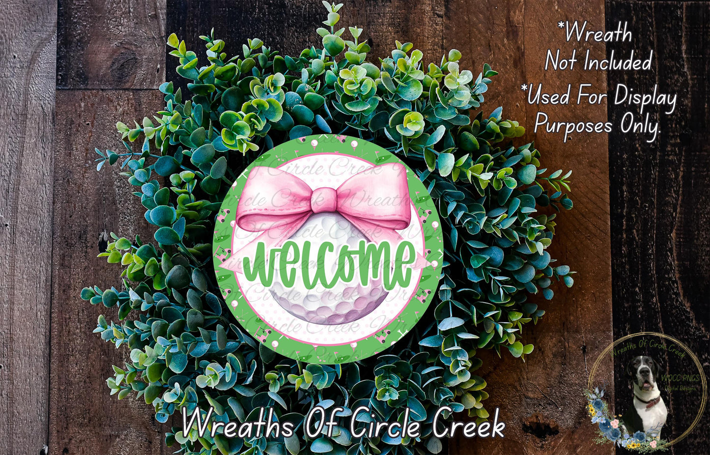 a wreath made of green and white plants, with a pink bow and the word "Welcome" written on it. The wreath is displayed on a wooden surface, and there is a small dog in the bottom right corner of the image.