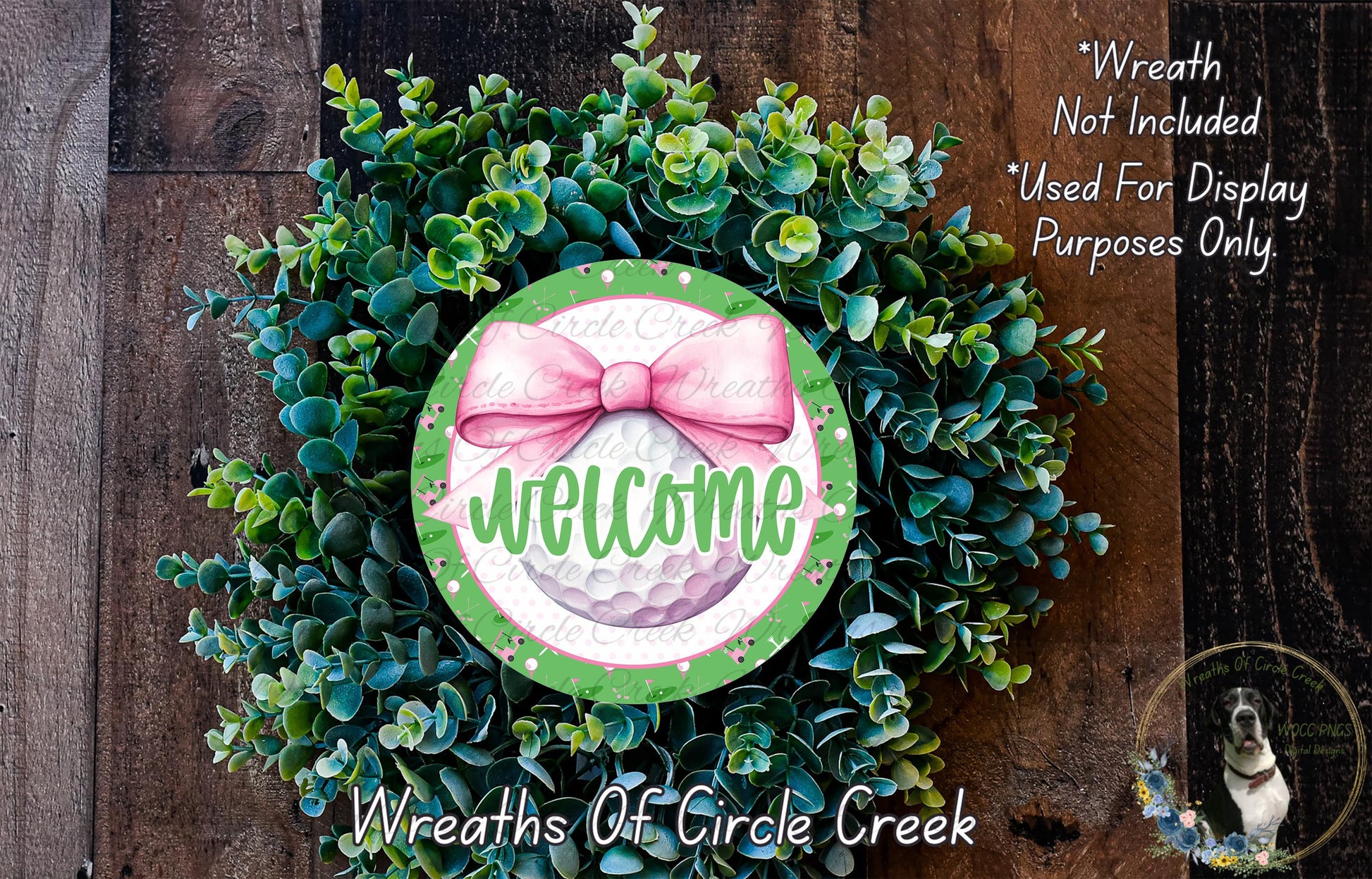 a wreath made of green and white plants, with a pink bow and the word "Welcome" written on it. The wreath is displayed on a wooden surface, and there is a small dog in the bottom right corner of the image.