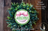 a wreath made of green and white plants, with a pink bow and the word "Welcome" written on it. The wreath is displayed on a wooden surface, and there is a small dog in the bottom right corner of the image.