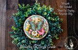 a wreath with a butterfly and flowers, surrounded by greenery. The wreath is placed on a wooden surface, and there is a small dog in the bottom right corner of the image.