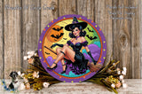 A round wall decoration featuring a woman dressed as a witch, with a Halloween-themed background and bats flying around. The decoration is surrounded by flowers and placed on a wooden surface.