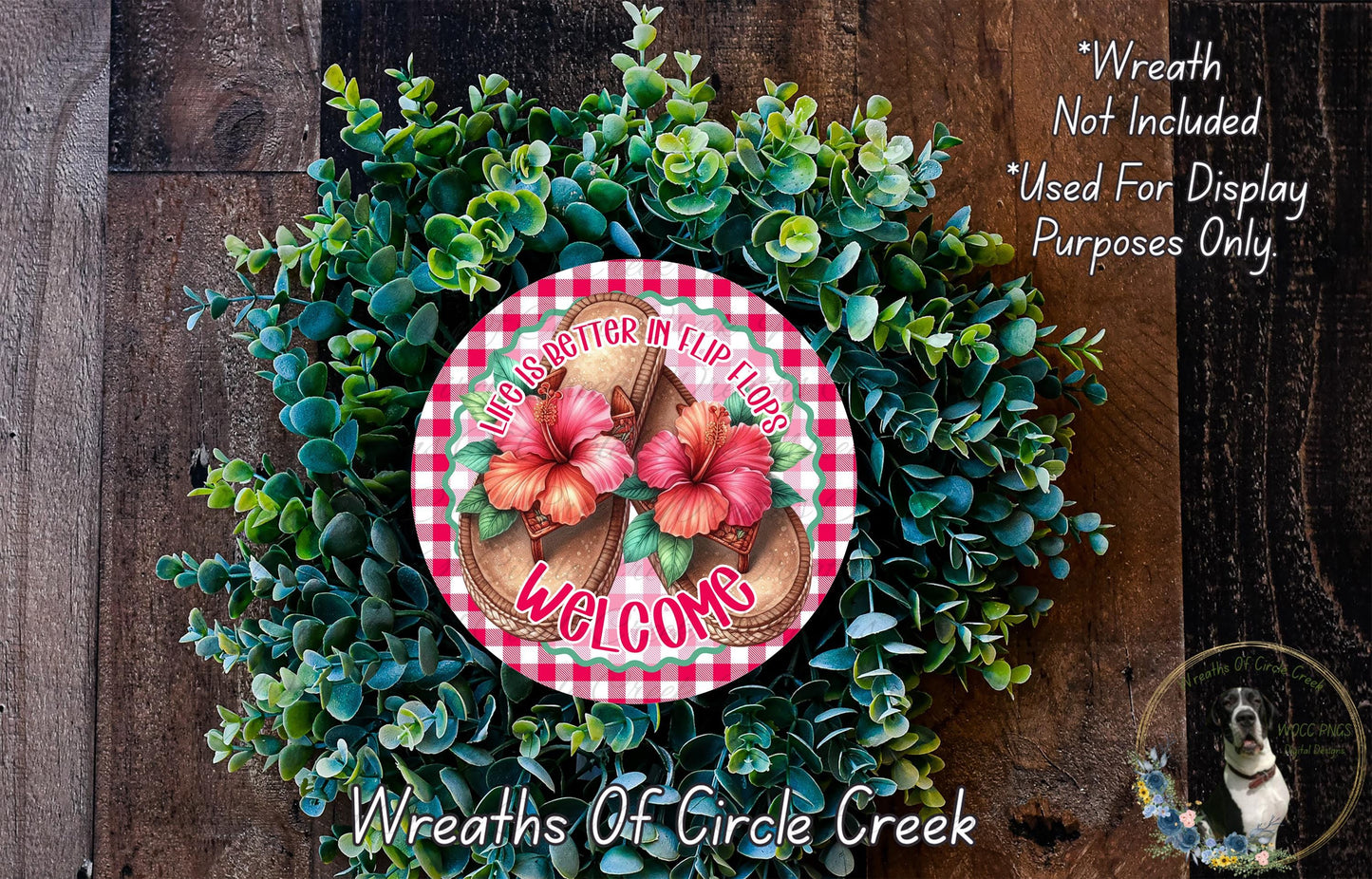a wreath made of green foliage and pink flowers, with a red and white circular sign that reads "Welcome" in the center. The wreath is displayed on a wooden surface, and there is a small dog in the bottom right corner of the image.