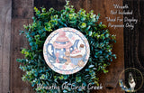 A wreath made of green foliage and featuring a decorative plate with a tea set design, including a teapot, cup, and saucer.