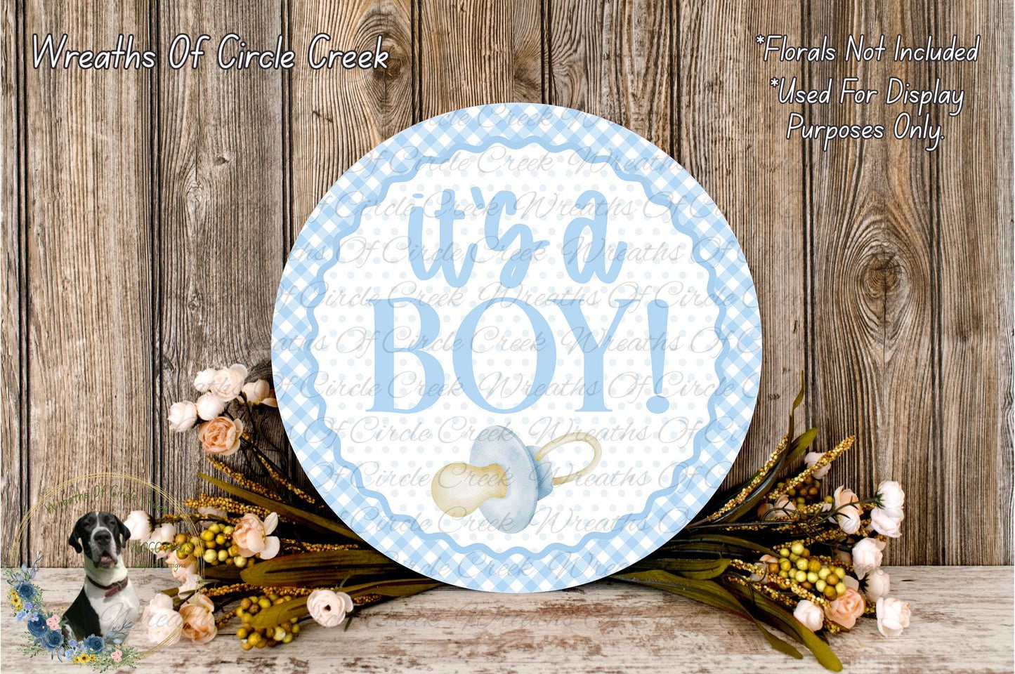 a round sign with the text "It's a BOY!" surrounded by a wreath of flowers and greenery, placed on a wooden surface.