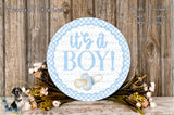 a round sign with the text "It's a BOY!" surrounded by a wreath of flowers and greenery, placed on a wooden surface.