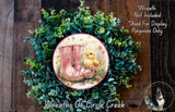 A wreath made of green leaves and flowers, with a pink boot and a yellow chick on it, placed on a wooden surface.