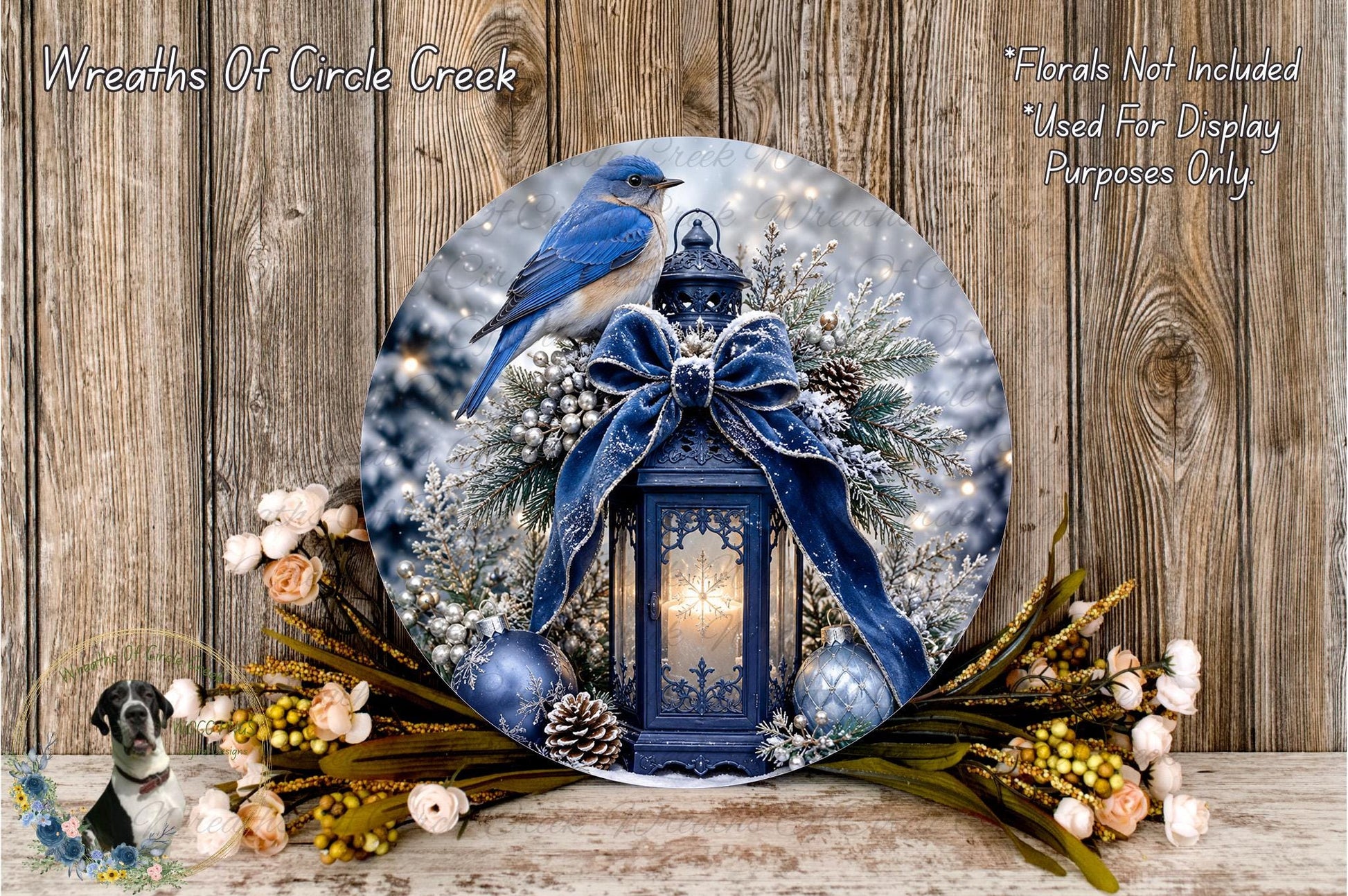 A blue bird perches on a wreath adorned with a lantern, pine cones, and other natural elements, set against a snowy landscape.