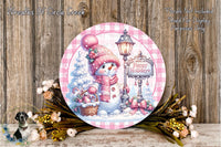 A round decorative plate featuring a snowman, a lamp post, and a sign that says "Happy Holidays" against a snowy background. The plate is surrounded by flowers and placed on a wooden surface.