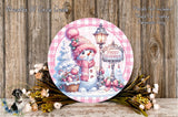A round decorative plate featuring a snowman, a lamp post, and a sign that says "Happy Holidays" against a snowy background. The plate is surrounded by flowers and placed on a wooden surface.