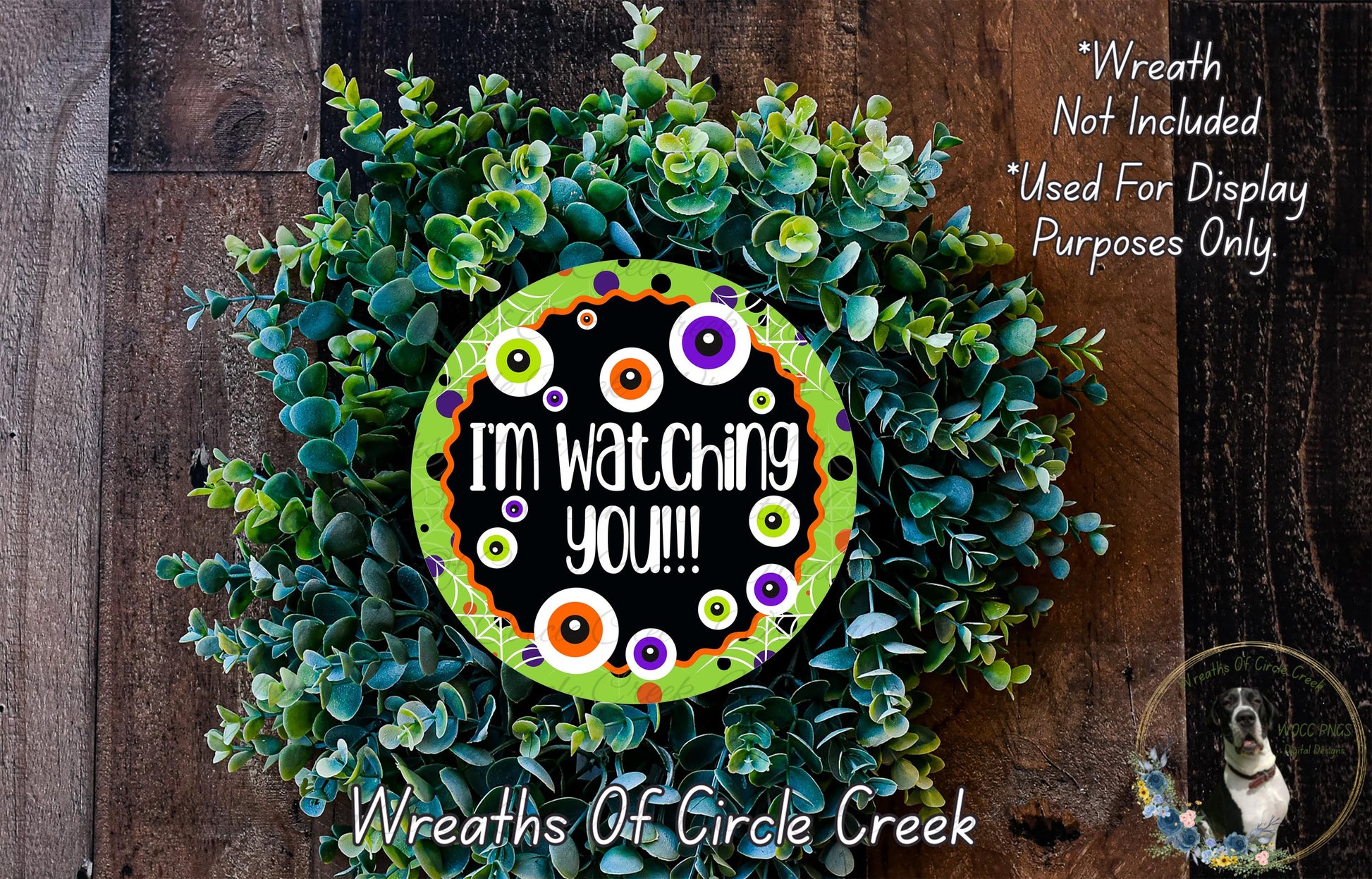 The image is a wreath made of green foliage, with a black and orange circular sign in the center that says "I'm watching you!". The wreath is displayed on a wooden surface, and there is a small dog in the bottom right corner.