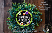 The image is a wreath made of green foliage, with a black and orange circular sign in the center that says "I'm watching you!". The wreath is displayed on a wooden surface, and there is a small dog in the bottom right corner.