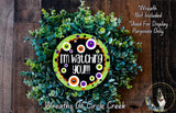 The image is a wreath made of green foliage, with a black and orange circular sign in the center that says "I'm watching you!". The wreath is displayed on a wooden surface, and there is a small dog in the bottom right corner.