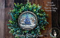A wreath made of evergreen branches and a circular design featuring a winter scene with a Christmas tree, houses, and presents. The wreath is displayed on a wooden surface, and there is a text overlay that reads "Wreath Not Included * Used For Display Purposes Only".