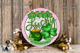 A decorative sign featuring the word "Lucky" surrounded by a pot of gold coins, a clover, and other festive elements, placed on a wooden surface with flowers and greenery.
