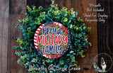 a wreath made of green leaves and a circular sign with the text "Proud Military Family" in the center. The wreath is displayed on a wooden surface, and there is a small dog in the bottom right corner of the image.