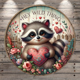 A raccoon holding a heart-shaped object is surrounded by a floral arrangement and a wooden fence, with the text "Hey Wild Thing" above the raccoon.