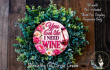 A wreath made of green leaves and pink flowers, with a pink sign that says "You look like I need wine".