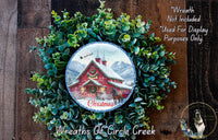 A Christmas wreath with a red barn and snowy mountain backdrop, surrounded by greenery. The wreath has a Santa Claus sleigh flying over the barn.