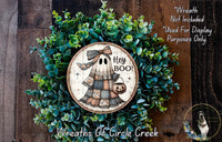 a Halloween-themed wreath with a ghost holding a "Hey Boo!" sign, surrounded by green foliage. The wreath is displayed on a wooden surface, and there is a small dog visible in the bottom right corner.