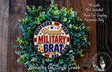 a wreath made of green leaves and a circular emblem with the text "Proud MILITARY BRAT" on it. The wreath is displayed on a wooden surface, and there is a small dog in the bottom right corner of the image.