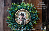 A wreath with a cat sitting in a window, surrounded by greenery, and a sign that says "Wreath Not Included Used For Display Purposes Only".
