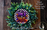 a wreath with the text "Trick or Treat" on it, surrounded by green foliage. The wreath is placed on a wooden surface, and there is a small dog in the bottom right corner of the image.
