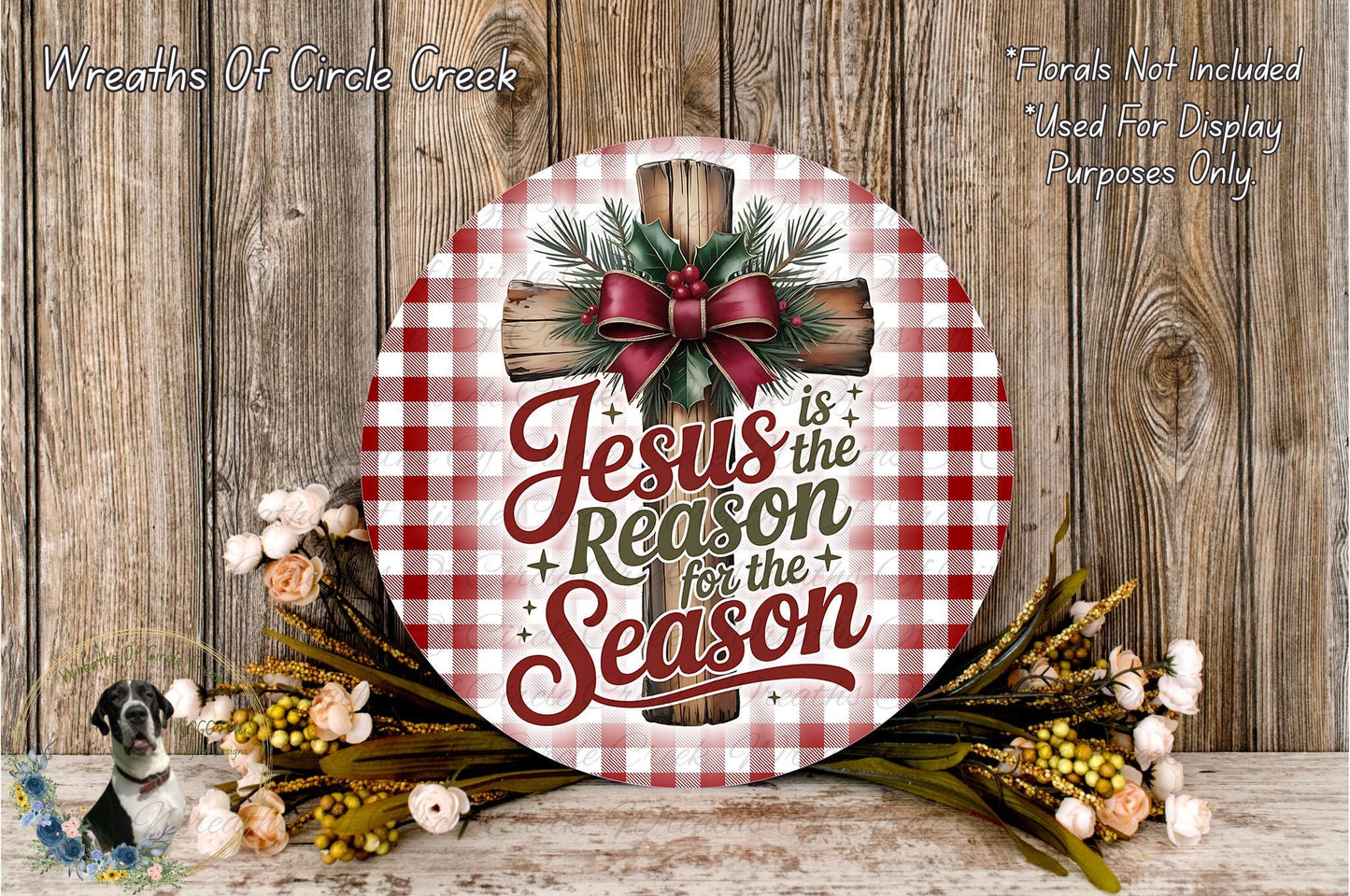 A round wooden sign with a red and white checkered pattern, featuring a cross and the text "Jesus is the reason for the season". The sign is surrounded by a wreath of flowers and greenery, and there is a wooden wall in the background.