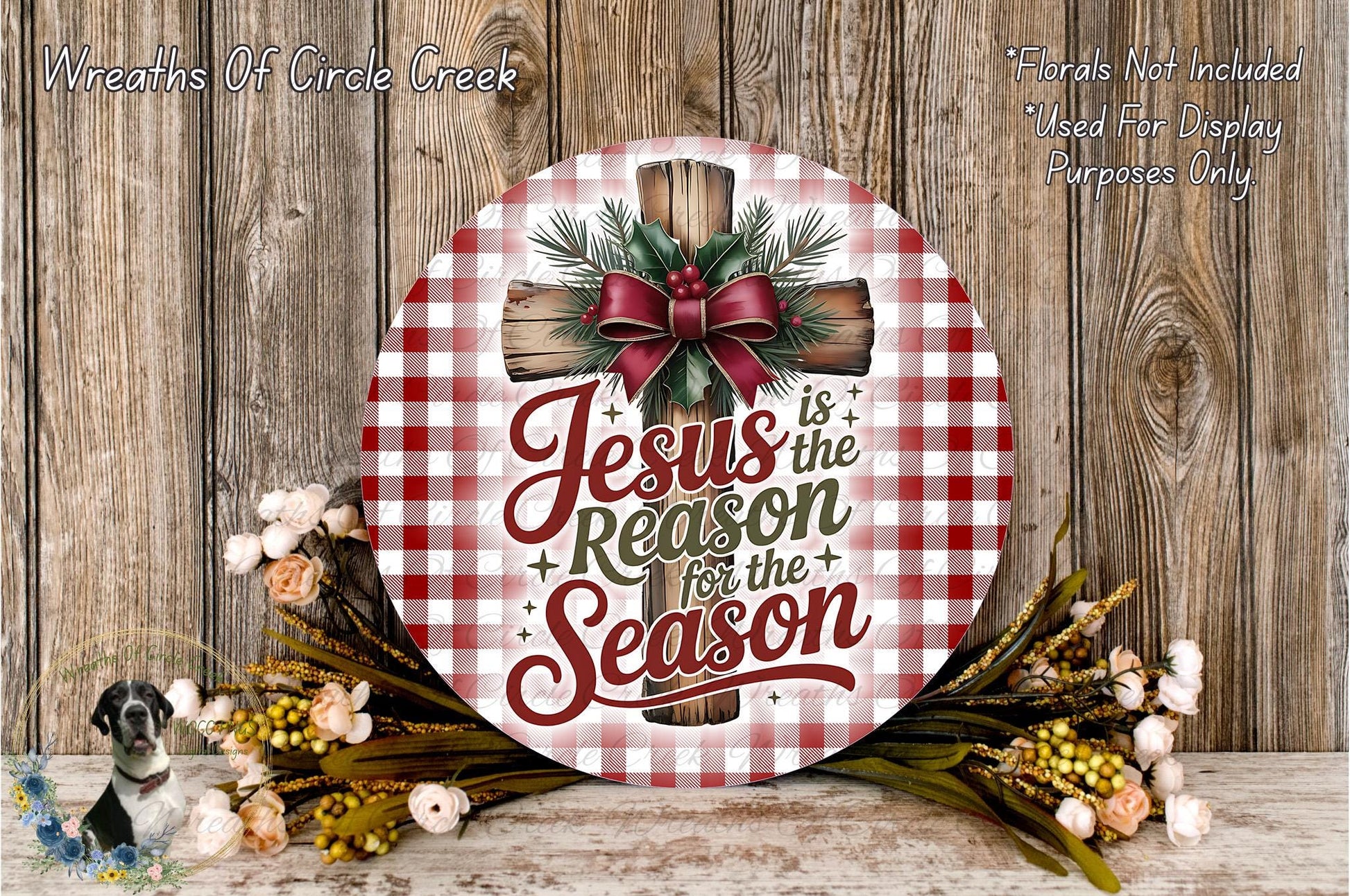 A round wooden sign with a red and white checkered pattern, featuring a cross and the text "Jesus is the reason for the season". The sign is surrounded by a wreath of flowers and greenery, and there is a wooden wall in the background.