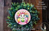 a wreath with a bunny wearing a pink bow and surrounded by green leaves and flowers. The wreath is placed on a wooden surface.