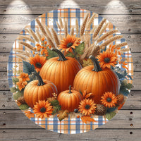 A circular arrangement of three pumpkins, two orange and one yellow, surrounded by sunflowers, pine cones, and other fall-themed elements against a plaid background.