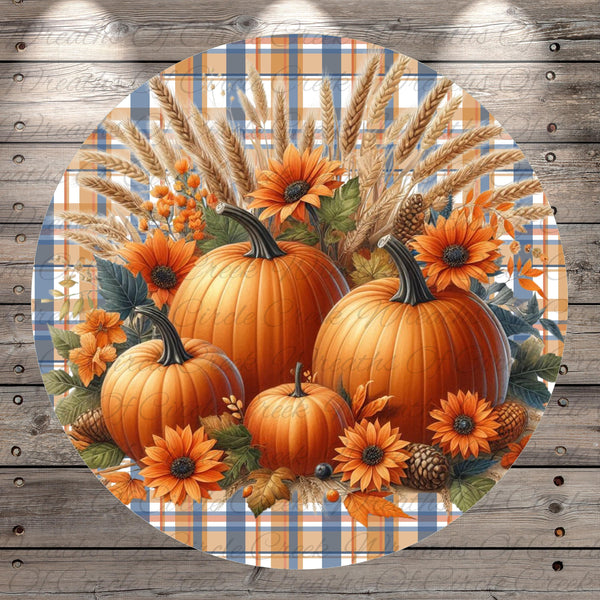 A circular arrangement of three pumpkins, two orange and one yellow, surrounded by sunflowers, pine cones, and other fall-themed elements against a plaid background.