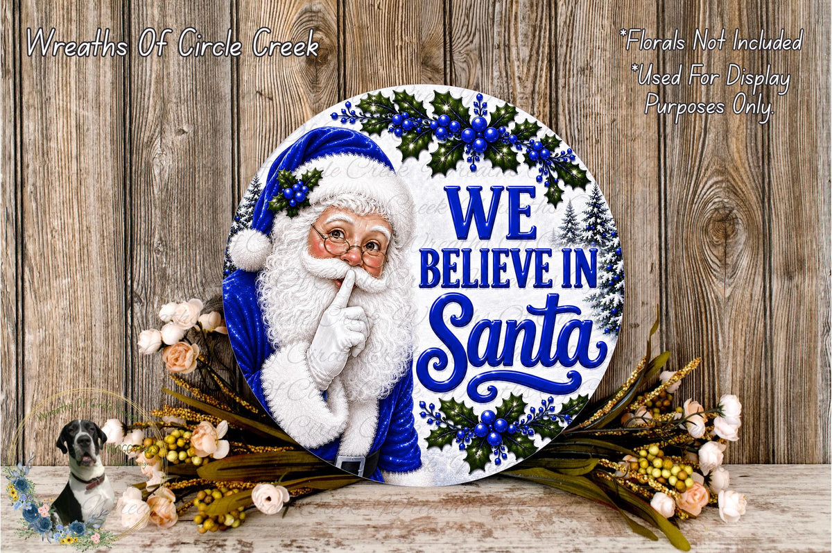 A round wooden sign with a Santa Claus image and the text "We Believe in Santa" is displayed on a wooden surface, surrounded by flowers.