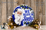 A round wooden sign with a Santa Claus image and the text "We Believe in Santa" is displayed on a wooden surface, surrounded by flowers.