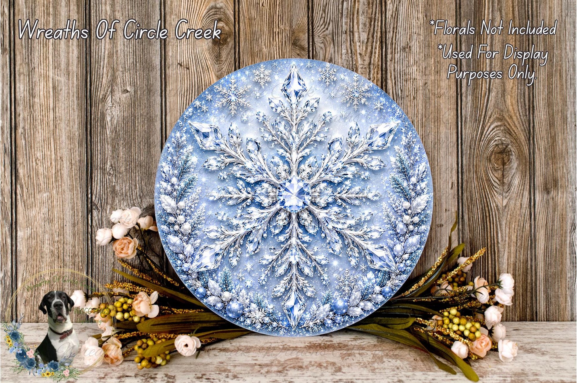 A large, round wreath made of blue and white materials, resembling a snowflake, is displayed on a wooden surface. The wreath is surrounded by a variety of flowers and greenery, creating a festive and natural setting.