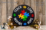 A black sign with colorful puzzle pieces and the words "Teach Love Inspire Hope" written on it, surrounded by flowers and greenery.