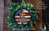 A wreath made of green leaves and a wooden cross, with the text "God & Country" displayed on it. The wreath is set against a wooden background, and there is a small dog in the bottom right corner.