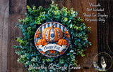 a wreath made of various fall-themed elements, including a pumpkin, greenery, and a banner that says &quot;Happy Fall&quot;. The wreath is displayed on a wooden surface, and there is a small dog in the bottom right corner of the image.