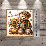 A teddy bear wearing a plaid hat and overalls, sitting next to a sunflower, pumpkins, and a wooden sign that says "Welcome".