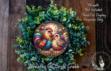 A colorful wreath featuring two vibrant roosters, surrounded by lush greenery, with the word "LOVE" prominently displayed. The wreath is set against a rustic wooden background, and there is a small dog visible in the bottom right corner.